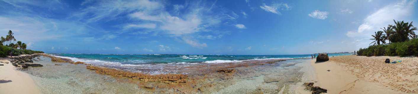 Panoramic beach scene showing turquoise Caribbean ocean water with visible coral reef formations, golden sand beach, palm trees, and blue sky with white clouds in San Andrés, Colombia