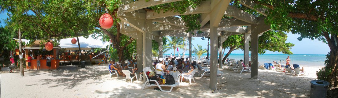 People sitting on white lounge chairs under a wooden beach pavilion with green vines, red lanterns hanging from trees, a beach bar with orange walls, white sand, and turquoise Caribbean ocean in the background