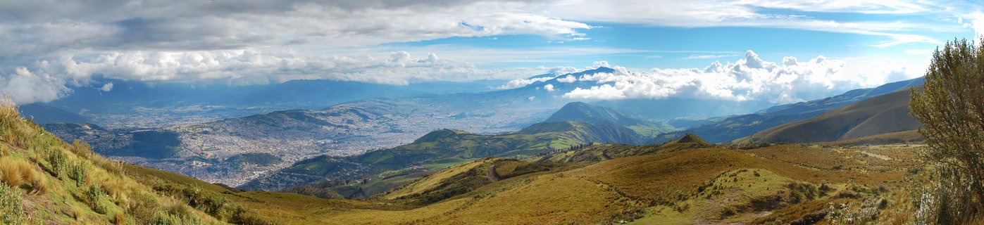 Wide panoramic landscape showing Quito city in a valley surrounded by green and brown mountain hillsides with distant volcanic peaks and cloudy blue sky