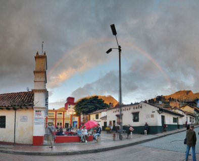 Street scene in Bogotá's La Candelaria neighborhood showing a rainbow over colonial white buildings with red tile roofs, people sitting on benches, and mountains in the background under cloudy skies