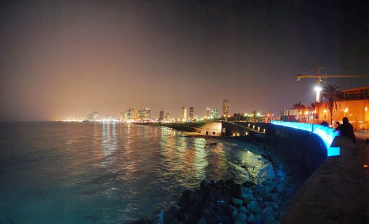 Nighttime coastal cityscape showing illuminated buildings along a shoreline with water reflections and a blue-lit seawall promenade