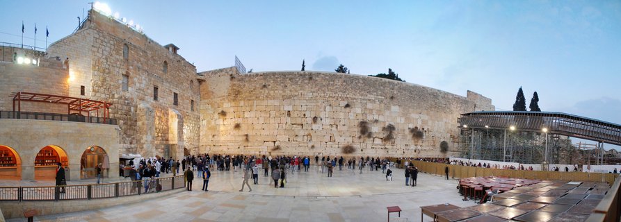 Panoramic view of the Western Wall in Jerusalem showing the ancient stone wall with crowds of people in the plaza, Israeli flags visible on buildings, and the Mughrabi Gate ramp on the right side