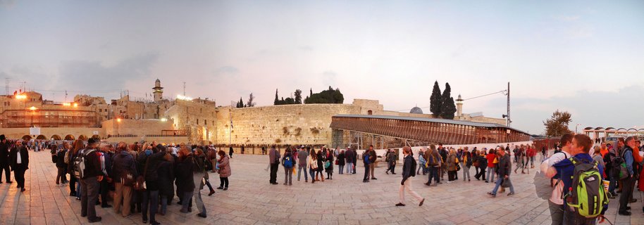 Panoramic view of the Western Wall plaza in Jerusalem at dusk with numerous visitors walking and praying in front of the illuminated ancient limestone wall