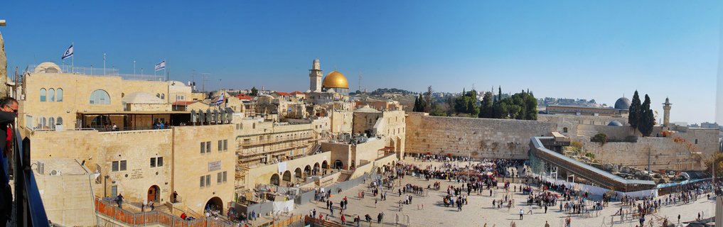 Panoramic view of Western Wall Plaza in Jerusalem with Dome of the Rock golden dome visible above ancient stone buildings under clear blue sky with crowds of people in the courtyard