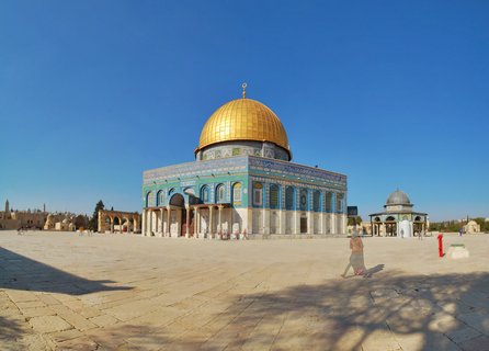 The Dome of the Rock with its golden dome and blue-tiled walls stands on a stone plaza under a clear blue sky in Jerusalem, with people walking in the foreground