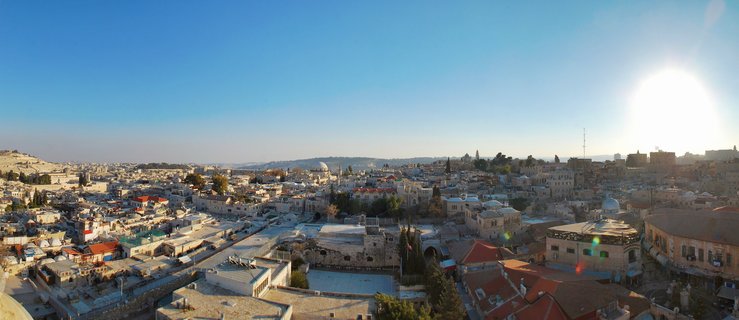 Aerial panoramic view of Jerusalem Old City showing limestone buildings with red-tiled roofs, Church of the Holy Sepulchre domes, bell tower, and clear blue sky with bright sun