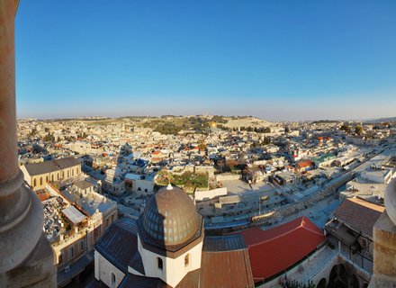 Panoramic cityscape of Jerusalem showing densely packed limestone buildings with multiple domes including the golden Dome of the Rock, viewed from an elevated position with stone columns in the foreground under a clear blue sky