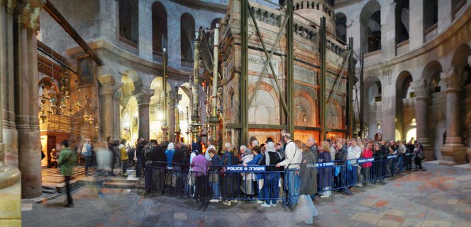 Interior view of the Church of the Holy Sepulchre showing pilgrims queuing behind blue police barriers toward the Aedicule structure with scaffolding, surrounded by stone columns and arches