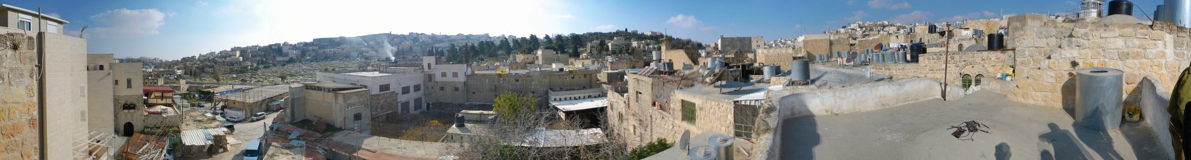 Wide panoramic photograph showing densely packed beige stone buildings of Hebron, Palestine, with flat rooftops, water tanks, and hills in the background under a blue sky with scattered clouds