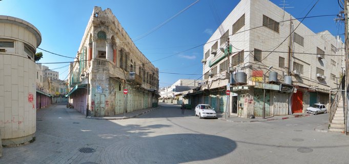 Street view of traditional Palestinian stone buildings in Hebron with closed shop shutters, parked cars, and overhead power lines under clear blue sky