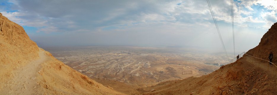 Wide desert panorama showing rocky beige cliffs framing a vast valley landscape with distant mountains under cloudy blue sky, two hikers visible on right-side trail with railings