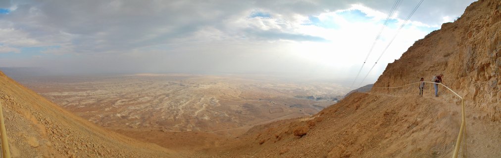 Panoramic view from Masada showing two people hiking on a winding path along a rocky cliffside, with vast desert valley and distant mountains under cloudy sky