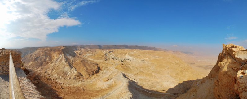 Panoramic desert landscape showing Masada fortress ruins with rocky hills, winding roads, and blue sky with scattered clouds in Israel's Judean Desert near the Dead Sea