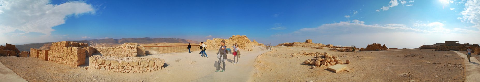 Wide panoramic view of ancient stone ruins at Masada fortress with tourists walking on pathways through the archaeological site in the Judean Desert, Israel, under bright blue sky with scattered clouds