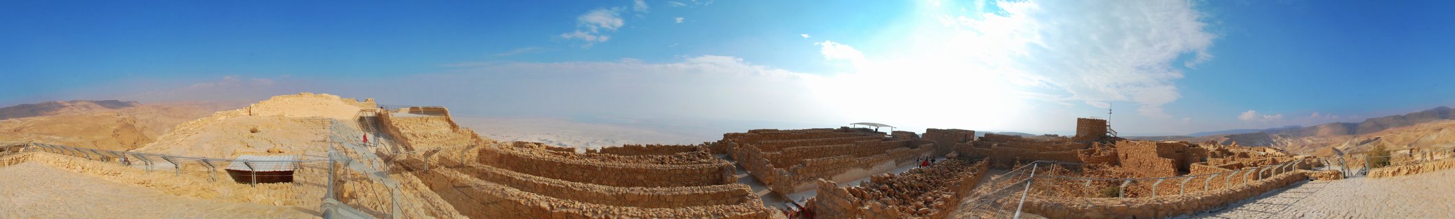 Panoramic view of Masada fortress ruins with ancient stone walls and archaeological excavations under blue sky, Dead Sea visible in the distance