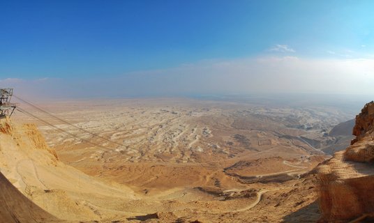 Panoramic view of Masada archaeological site in the Judean Desert showing ancient ruins, cable car lines, winding desert paths, and rocky cliffs under a clear blue sky near the Dead Sea in Israel
