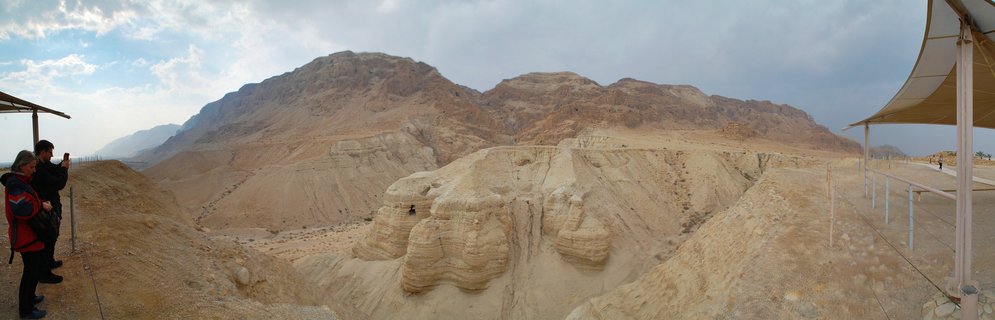 Panoramic desert landscape showing beige layered rock formations with mountains in the background, tourist viewing areas with white shade canopies on both sides, and two visitors on the left
