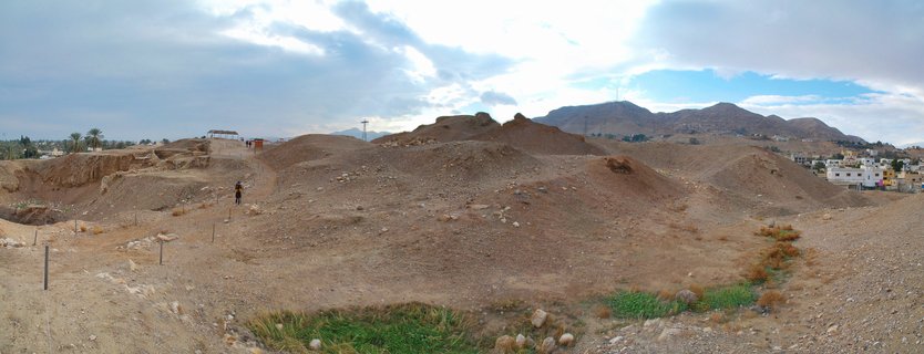 Panoramic view of an archaeological excavation site in the Dead Sea desert with brown earth mounds, a walking visitor, distant mountains, and white buildings under cloudy sky