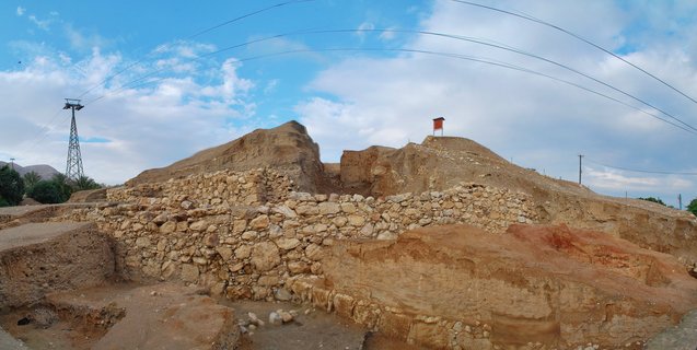 Ancient stone wall ruins at archaeological excavation site with earthen mounds, cable car tower, and blue sky with clouds in Israel