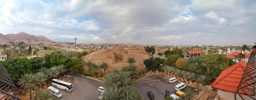 Panoramic aerial view of archaeological mound in Jericho with tourist buses, palm trees, red-roofed buildings, and desert mountains under cloudy sky