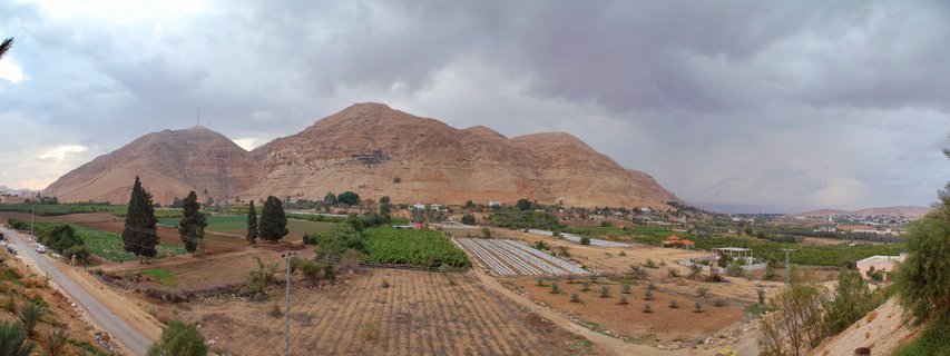 Wide landscape showing green agricultural fields and white greenhouse rows in a valley with large brown desert mountains under cloudy sky