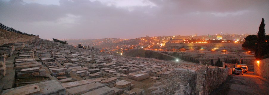Panoramic view of Mount of Olives Jewish Cemetery with rows of stone tombs in foreground, Jerusalem Old City and Dome of the Rock illuminated in the distance at twilight
