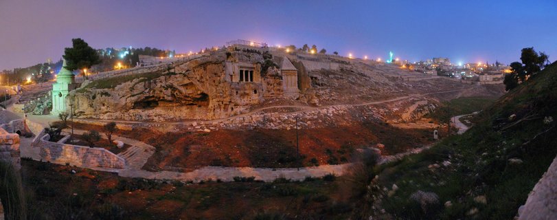 Ancient rock-cut tombs carved into limestone hillside on Mount of Olives in Jerusalem at twilight, illuminated by warm artificial lighting against deep blue evening sky, with winding paths in foreground and green-domed structure visible on left side