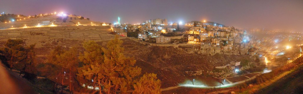 Nighttime panoramic photograph of Jerusalem showing illuminated hillside buildings, cemetery terraces on Mount of Olives, and scattered trees under a dark purple sky