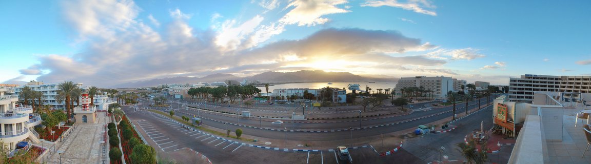 Panoramic aerial view of a coastal resort city at sunrise showing hotels, palm trees, empty roads with a large roundabout, Red Sea water, and mountains in the distance under a blue sky with scattered clouds