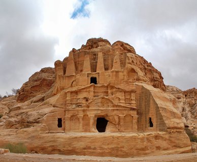 Ancient rock-cut tomb facade with multiple levels, columns, and triangular pediments carved into orange sandstone cliff under cloudy sky at Petra, Jordan