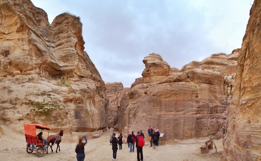 Wide view of the Siq canyon at Petra showing towering pink and orange sandstone cliffs on both sides with tourists walking on a dirt path and a horse-drawn carriage with orange canopy on the left under an overcast sky
