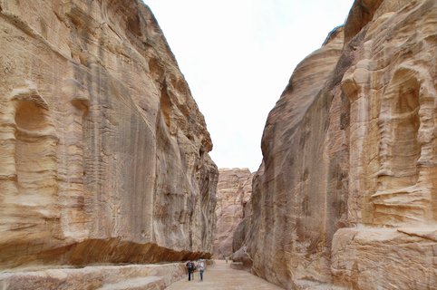 Two tourists walking on a pathway through a narrow sandstone canyon with towering rock walls on both sides at Petra archaeological site in Jordan