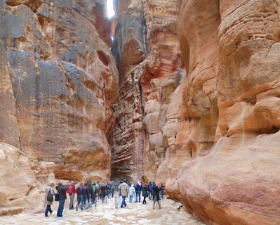 Group of tourists walking through narrow sandstone canyon with towering reddish-brown rock walls at Petra, Jordan