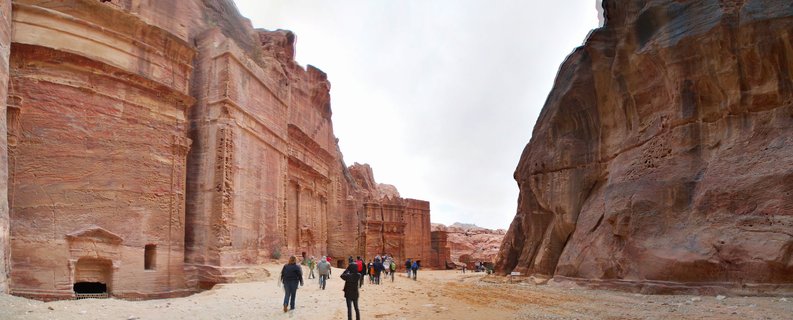 Tourists walking on a sandy path between massive rose-red rock-cut ancient facades carved into cliff walls at Petra archaeological site in Jordan