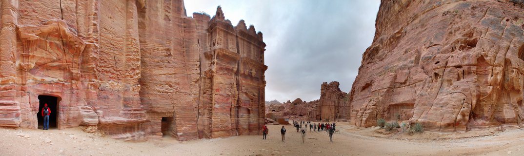 Wide view of ancient rock-cut tomb facades carved into red sandstone cliffs at Petra, Jordan, with tourists walking on sandy ground between the monuments under an overcast sky