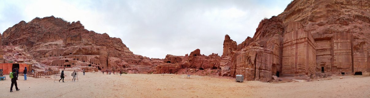 Wide view of ancient rock-cut tomb facades carved into red sandstone cliffs at Petra, Jordan, with tourists walking on sandy ground below under cloudy sky