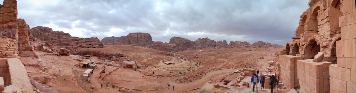 Wide panoramic photograph showing the ancient archaeological site of Petra in Jordan with carved sandstone ruins, rock-cut tombs, desert terrain, and tourists walking on pathways under cloudy skies