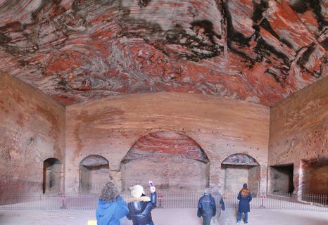 Tourists photograph the interior of a carved sandstone chamber at Petra, Jordan, featuring distinctive red and white striated rock formations and ancient arched niches.
