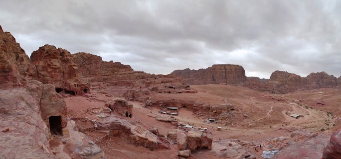 Wide panoramic photograph showing ancient rock-cut tomb facades carved into rose-red sandstone cliffs at Petra archaeological site in Jordan, with valley floor and overcast sky
