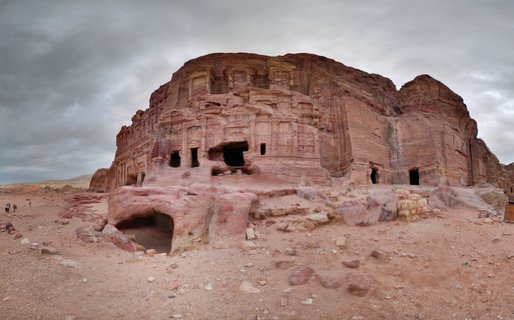 Ancient rock-cut tomb facades with classical columns and pediments carved into rose-red sandstone cliffs at Petra, Jordan, with scattered rocks in the desert foreground and tourists visible on the left side under a cloudy sky