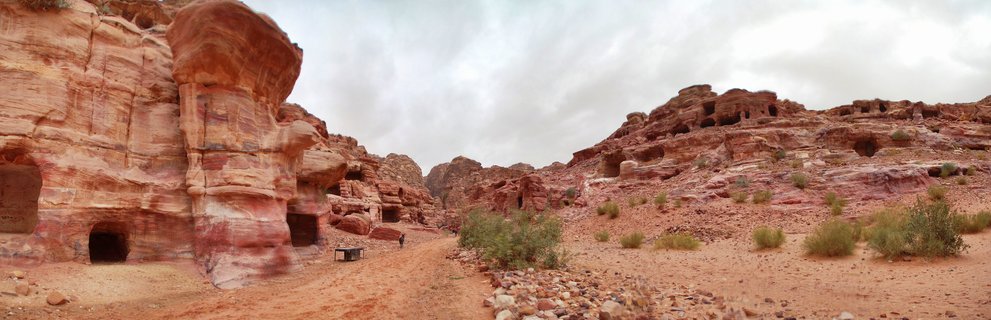 Ancient rock-cut tomb facades carved into red sandstone cliffs at Petra archaeological site in Jordan with desert path and sparse vegetation