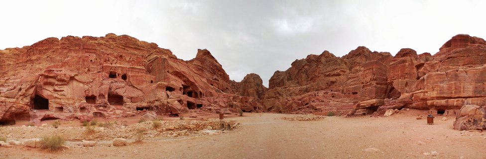 Wide view of ancient rock-cut tomb facades carved into red sandstone cliffs at Petra archaeological site in Jordan with desert pathway and overcast sky