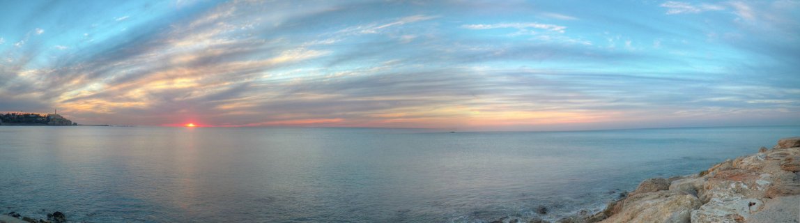 Panoramic seascape showing sunset over calm Mediterranean waters with rocky coastline in foreground and distant city silhouette on the horizon