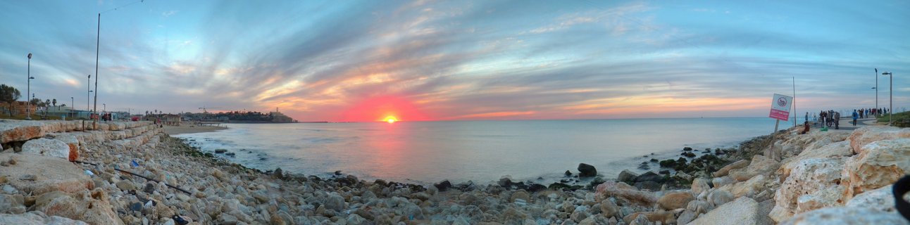 Panoramic sunset view over the Mediterranean Sea from Tel Aviv's rocky coastline with people walking along the promenade and street lamps visible