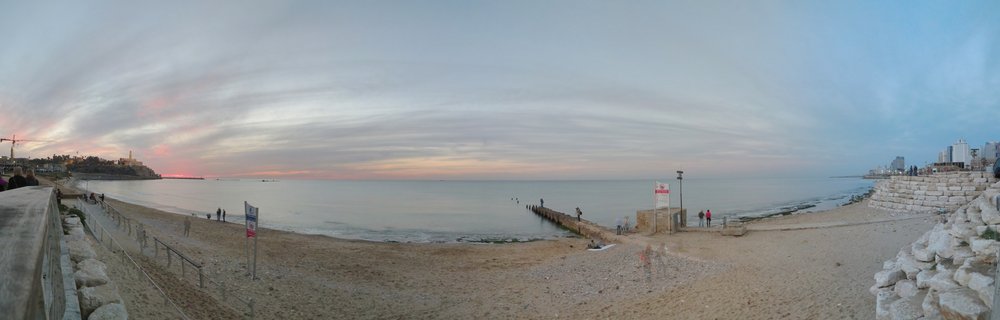 Wide panoramic beach scene showing Tel Aviv coastline at sunset with sandy shore, calm Mediterranean Sea, pier, stone walls, people walking, historic buildings on left and modern city skyline on right under colorful sky