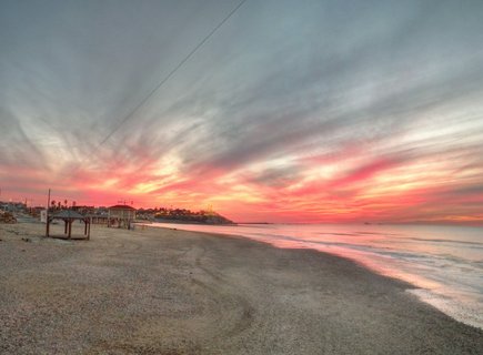 Panoramic beach scene at sunset showing sandy shore with wooden gazebos, calm Mediterranean Sea with pink sky reflections, and dramatic orange and pink clouds over distant Tel Aviv cityscape