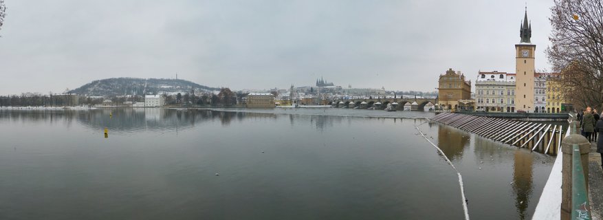 Wide panoramic view of Prague showing the Vltava River with Charles Bridge and Prague Castle with cathedral spires visible on a hill in the background during winter with snow on buildings and bare trees along an overcast sky