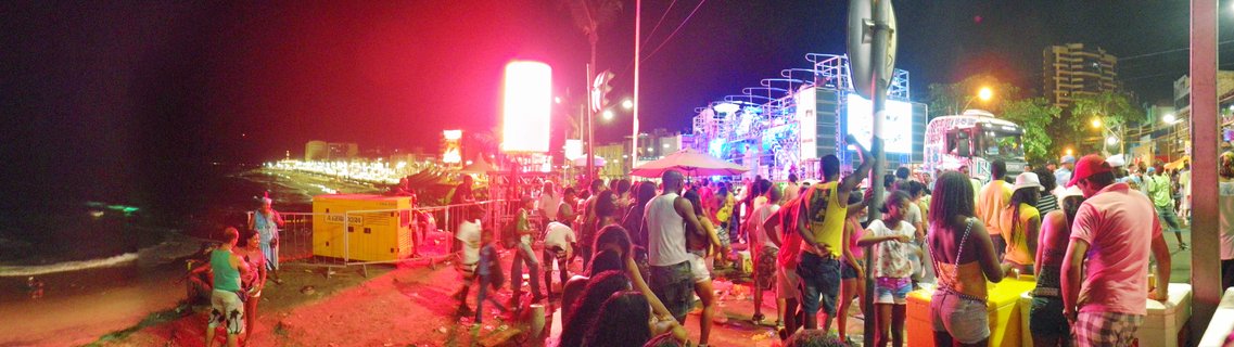 Nighttime panoramic view of a crowd at a Carnival street party in Salvador, Brazil, with a lit stage, red lighting, and ocean visible on the left side