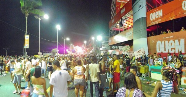 Nighttime street scene showing large crowd at Carnival celebration in Salvador Brazil with multi-level orange camarote structure, palm trees, and street lighting