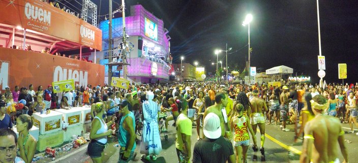 Nighttime street scene of Brazilian Carnival in Salvador with large crowd of people celebrating, orange VIP box structure with QUEM branding, illuminated pink building, and street lights